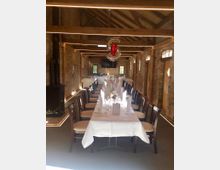 Langer, dekorierter Tisch in einem rustikalen Raum mit Backsteinwänden, Holzdecke und sichtbaren Holzbalken. Im Hintergrund sind Fenster, ein Kaminofen und ein Fernseher sichtbar.