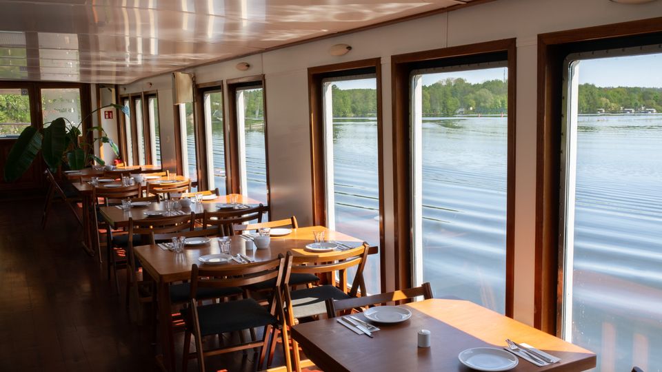 Innenraum eines Restaurants auf einem Boot mit Holzböden und -möbeln. Große Fenster auf der rechten Seite bieten einen Ausblick auf ein ruhiges Gewässer und grüne Uferlandschaft.