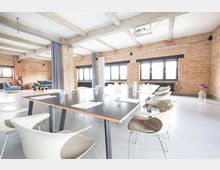 Moderner Konferenzraum in einem Loft mit Backsteinwänden, einer langen dunklen Tischplatte, weißen Stühlen und großen Fenstern, die viel Tageslicht einlassen. Der Raum ist schlicht eingerichtet mit sichtbaren Balken an der Decke und dekorativen Elementen wie Blumen in einer Glasvase.