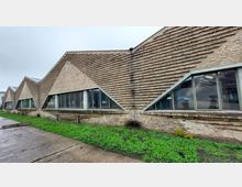 Backstein-Gebäude mit schräg verlaufenden Ziegelreihen und großen, dreieckigen Fenstern, die in regelmäßigen Abständen angeordnet sind. Im Vordergrund eine Asphaltfläche und Grünflächen, im Hintergrund bewölkter Himmel.