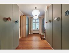 Schmaler Raum mit Holzboden, grünen Einbauschränken auf beiden Seiten und verspielten, bunten Türknöpfen. Am Ende des Raums befindet sich ein Fenster mit Vorhängen, darunter ein Heizkörper, sowie ein Holzstuhl an der rechten Wand.