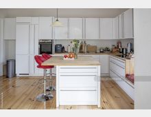 Helle, moderne Küche mit weißen Hochglanz-Einbauschränken, einer Arbeitsinsel mit Holzoberfläche und roten Barhockern. Die Einrichtung umfasst eine eingebaute Spüle, Elektrogeräte und ein offenes Regal; der Boden besteht aus hellem Holz.