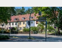 Straßenszene mit einem kleinen Wohngebiet, bestehend aus Reihenhäusern mit roten Ziegeldächern und bunten Fassaden in Gelb, Blau und Weiß. Im Vordergrund befinden sich Straßenschilder der Kreuzung „Onkel-Tom-Straße“ und „Riemeisterstraße“ sowie Verkehrszeichen und ein gelber Briefkasten, umgeben von Bäumen.