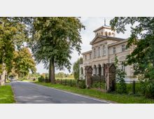 Historisches Herrenhaus im klassizistischen Stil mit heller Fassade, Rundbögen und einem Turmaufsatz, umgeben von einem Metallzaun und einer grünen Landschaft. Eine von Bäumen gesäumte Landstraße führt links am Gebäude vorbei.