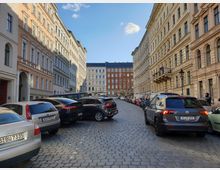 Straße mit gepflastertem Belag, gesäumt von mehrstöckigen Gründerzeitgebäuden mit hellen Fassaden. Am Straßenrand stehen parkende Autos, der Himmel ist klar mit wenigen Wolken.