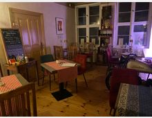 Gemütlicher Gastraum eines kleinen Restaurants mit Holzboden, karierten Tischdecken, bunten Stühlen und einer Kreidetafel mit Speisekarte neben einer Holztür. Im Hintergrund stehen hohe Fenster, ein Regal mit Weinflaschen und ein Thekentisch; rechts ist ein dekoratives Motorrad im Innenraum zu sehen.