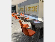 Außenbereich eines Cafés mit orangefarbenen Plastikstühlen und steinernen Tischen auf einem gepflasterten Boden. Im Hintergrund eine große Glasfront mit gelbem Schriftzug „AL CATZONE“.
