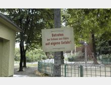 Ein Schild mit der Aufschrift „Betreten bei Schnee und Glätte auf eigene Gefahr!“ hängt an einem Metallpfosten in einem Park. Im Hintergrund sind ein Klettergerüst, ein Zaun, Bäume und ein etwaiges Gebäude zu sehen.