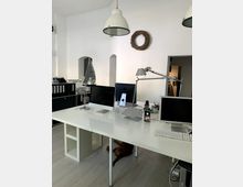 Ein Arbeitsraum mit weiß gestrichenen Wänden, einem großen weißen Tisch mit mehreren Computermonitoren, Schreibtischlampen und Bürobedarf. Im Hintergrund befinden sich Regale mit Aktenordnern, eine schlichte Dekoration aus einem Kranz an der Wand und moderne Hängelampen an der Decke.