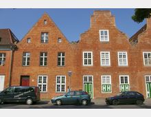 Backsteingebäude mit Satteldach und geschwungenem Giebel im traditionellen Baustil, mehrere weiße Fenster und grüne Türen mit Ziermustern. Im Vordergrund parken Autos entlang der Straße bei klarem Himmel.