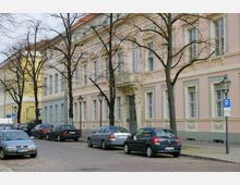 Straßenszene mit gepflasterter Fahrbahn, parkenden Autos und einem historischen Gebäude mit rosa-weiß gestrichener Fassade, großen Fenstern und detaillierten architektonischen Verzierungen. Im Vordergrund stehen Bäume und Laternen, die den Gehweg säumen.