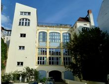 Wohngebäude im Jugendstil mit cremefarbener Fassade, großen Rundbogenfenstern und goldenen Ornamenten, umgeben von Bäumen und geparkten Autos im Vordergrund. Im oberen Bereich des Gebäudes befindet sich eine Dachterrasse.