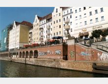 Flussufer mit mehrstöckigen historischen Gebäuden im Hintergrund, die weiße und gelbe Fassaden haben; entlang des Ufers eine Backsteinmauer mit Bögen, teils mit Graffiti bedeckt, und eine erhöhte Terrasse mit Schirmen und Sitzbereichen.