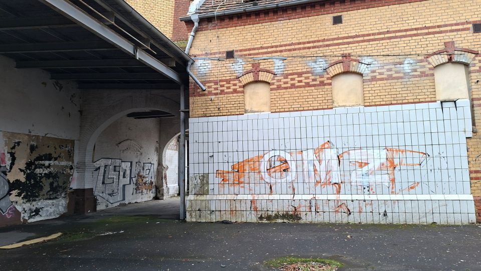 Überdachter Durchgang an einem alten Backsteingebäude mit zugemauerten Rundbogenfenstern und einem unteren Wandbereich aus weißen Fliesen. Die Wände sind teilweise abblätternd und mit Graffiti versehen; der Boden ist asphaltierter Hofbereich.