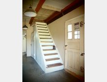 Ein rustikaler Innenraum mit einer Holztreppe, die mit weißen Wangen und abgenutzten Holzstufen gestaltet ist. Rechts daneben befindet sich eine alte Holztür mit Glasscheiben und Schnappschloss, während an der Decke Holzträger und Hängelampen sichtbar sind.