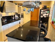 Moderne Küche mit hellen Einbauschränken, schwarzer Granitarbeitsplatte und zentraler Kochinsel mit schwarzen Barhockern. Im Hintergrund ein Holzrahmentor, ein schwarzer Kühlschrank mit Magneten und eine Fensterbank mit Pflanzen.