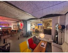 Industriewohnraum mit Gewölbedecke aus weiß gestrichenen Ziegeln, eingerichtet mit verschiedenen Sitzmöbeln, darunter bunte Stühle und Sofas. Auffällige Elemente sind ein großer Studioscheinwerfer, Neon-Schriftzüge an den Wänden und hängende Lampen; der Raum hat einen rustikalen und loftartigen Charakter.