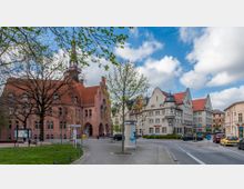 Straßenszene mit historischen Gebäuden: Links ein großes Backsteingebäude mit roten Dachziegeln und einem Bogenportal, rechts mehrere helle Gebäude mit Giebeln und roten Dächern. Im Vordergrund ein kleiner Platz mit Bäumen, Straßenschildern und parkenden Autos.