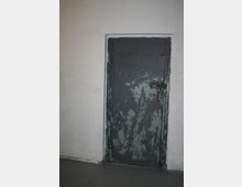 Eine dunkelgraue Metalltür mit sichtbaren Abblätterungen und Rostspuren, eingebaut in eine weiße, schlichte Wand. Die Tür ist mit Graffiti beschmiert und wirkt stark abgenutzt.