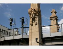 Ein steinernes Brückenelement mit einer dekorativen Frauenkopf-Skulptur und verzierten Säulen, flankiert von Metallgeländern und Ampelstrukturen im Hintergrund. Der Himmel ist blau mit leichten Wolken.