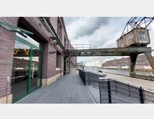 Backstein-Industriegebäude an einem Kanal mit angrenzendem Gehweg. Im Vordergrund sind eine grüne Glastür, ein Metallgeländer und ein alter, rostiger Kran, im Hintergrund Tanks und weitere Backsteingebäude.