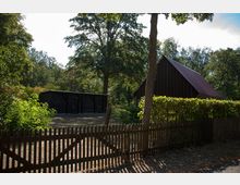 Holzzaun mit Tor im Vordergrund, dahinter eine umzäunte Gartenfläche mit einem Holzschuppen und einem Gebäude mit dunklem Holz und einem schrägen Dach. Umgeben von Bäumen und Sträuchern in einer ländlichen Umgebung.