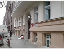 Straßenszene mit einem beigefarbenen Gebäude, das dekorative Stuckarbeiten und große Fenster aufweist, sowie einem Eingang mit einem verzierten Holztürrahmen. Im Vordergrund stehen ein geparktes Auto, ein Fahrrad und zwei Personen auf dem Gehweg neben einem Wohnmobil.