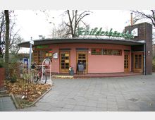 Ein kleines Restaurant mit rosa Fassade und Holzfenstern, über dem Eingang befindet sich ein großes grünes Neonschild mit der Aufschrift "Schildenhäuschen". Davor stehen ein Fahrradständer sowie ein Werbeschild, und links ist ein Bereich mit herbstlichem Laub und Pflanzen erkennbar.