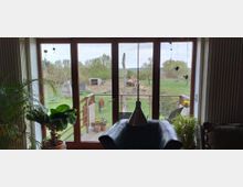 Ein Wohnzimmer mit großen Glastüren, die auf einen Balkon und eine weitläufige grüne Wiese führen. Im Außenbereich sind Bäume, ein Holzzaun, eine Kuh sowie eine kleine Hütte sichtbar; im Innenbereich befinden sich Pflanzen und ein Sessel.