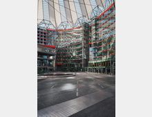 Innenhof des Sony Centers in Berlin mit moderner Glas- und Stahlarchitektur, roten Akzenten an den Gebäuden und einer markanten, zeltartigen Überdachung. Der Boden besteht aus dunklem Pflaster und Metallplatten, im Zentrum befindet sich ein runder Brunnen.