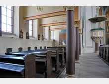 Innenraum einer Kirche mit Holzbänken, bemalten Säulen und verzierten Kapitellen. An der Wand befinden sich bunte Glasfenster und Kreuzwegstationen, rechts eine Kanzel, im Hintergrund ein Altar mit einer religiösen Skulptur.