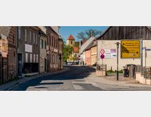 Quiet small-town street lined with older stucco and brick houses, curving toward a church tower with a red roof in the distance. A large yellow German direction sign and speed limit sign stand beside a wooden fence and barn-like building on the right.