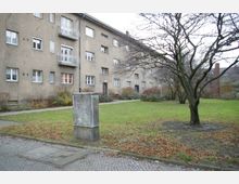 Wohnhaus in einem Innenhof mit einfachem Putzbau, mehreren Fenstern und Balkonen; im Vordergrund eine Grünfläche mit kahlen Bäumen sowie ein Stromkasten, umgeben von Gehwegen und Beeten.