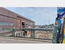 Bahnhof „Warschauer Straße“ in Berlin mit einer modernen, metallverkleideten Gebäudefassade, Fußgängern auf einer Brücke und sichtbaren Bahngleisen darunter, im Hintergrund ein Bogen einer Eisenbahnbrücke. Im Vordergrund ein mit Plakaten beklebter Mast.
