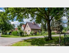 Wohngebiet mit Einfamilienhäusern, umgeben von Gärten und Bäumen; im Vordergrund ein großes grünes Grundstück mit einem Baum, neben einer Straßenlaterne und einem Straßenschild mit der Aufschrift "Wilhelm-Busch-Str."