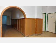 Flur mit einem braunen Rundbogen und holzvertäfelter Wand in der unteren Hälfte. Im Hintergrund führen zwei Türen und ein weiterer Durchgang in angrenzende Räume; die Türrahmen sind grün gestrichen.