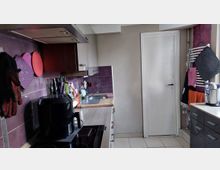 Schmale Küche mit L-förmiger Arbeitsfläche, Herd mit Dunstabzug und Spüle, flankiert von weißen und dunklen Schränken. Violette Fliesenrückwand und Wandpaneele, helle Bodenfliesen sowie eine weiße Tür am Ende des Raums; an der rechten Wand hängen Handtücher und Küchenutensilien.