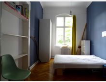Ein schlichter Raum mit Holzboden, einer dunkelblauen Wand und einer großen Fensterfront mit gelbem Vorhang. Der Raum enthält ein weißes Einzelbett, ein grüner Stuhl, ein Regal mit Büchern und andere kleinere Möbelstücke.