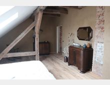 Dachgeschossraum mit rustikalem Charme, geprägt von sichtbaren Holzbalken, einer schrägen weißen Decke und freiliegenden Ziegelwänden. Der Raum ist mit Holzdielen, antiken Möbeln wie einer Kommode, einem Spiegel und dekorativen Vasen ausgestattet.