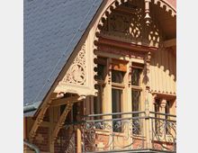 Fassade eines historischen Gebäudes mit detaillierten Holzschnitzereien und einem spitzwinkligen Satteldach aus Schiefer. Ein schmiedeeiserner Balkon und verzierte Fenster sowie kunstvolle Ornamentik prägen das Erscheinungsbild.