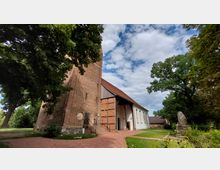 Ziegelkirche mit einem hohen Turm aus Backstein und einem anschließenden Kirchenschiff mit weiß verputzten Wänden und roten Dachziegeln, umgeben von Bäumen und einem gepflasterten Weg im Vordergrund. Der Himmel ist leicht bewölkt.
