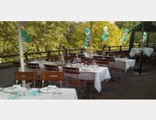 Eine ausgestattete Terrasse mit weiß gedeckten Tischen und Holzbänken, dekoriert mit weißen und türkisfarbenen Luftballons, umgeben von Bäumen im Hintergrund. Die Tische sind mit Gläsern, Besteck, Servietten und kleinen Dekorationen gestaltet.