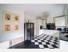 Helle Küche mit weißen Schränken, schwarzen Arbeitsplatten und einem schwarz-weiß karierten Boden. An der Wand hängen vier botanische Zeichnungen in goldenen Rahmen, und ein schwarzer Kühlschrank steht links neben einer Tür.