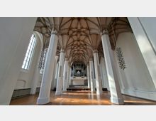 Innenraum einer großen Kirche mit hohen weißen Pfeilern, langen Seitenfenstern und einem reich gegliederten gotischen Kreuzrippengewölbe. Am Ende des Mittelschiffs steht eine Empore mit einer großen Orgel über einer erhöhten Bühne.