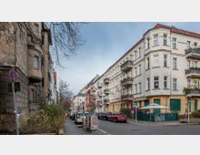 Straßenszene in einem Wohngebiet mit mehrstöckigen Altbauten, teils mit hellen Fassaden und Balkonen. Im Vordergrund sind parkende Autos, ein Verkehrsschild und graue Verteilerkästen zu sehen, umgeben von kahlen Bäumen und winterlicher Vegetation.