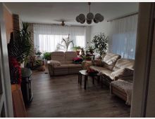 Helles Wohnzimmer mit großen Fenstern und transparenten Vorhängen, ausgestattet mit beigen Ledersofas, einem runden Couchtisch und zahlreichen Zimmerpflanzen. Der Boden besteht aus Holzlaminat, und von der Decke hängt eine moderne Lampe mit Kugel-Leuchten.