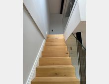 Innenaufnahme eines modernen Treppenhauses mit hellen Holzstufen, weißen Wänden und einem Glasgeländer. Die Treppe führt nach oben zu einem offenen Flur mit Glasbrüstung und sichtbarem dunklem Deckenbalken.