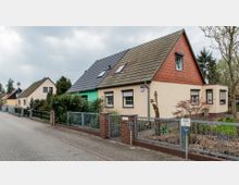 Straßenansicht mit Einfamilienhäusern mit Satteldächern, Vorgärten und Metallzäunen. Die Häuser sind unterschiedlich farbig gestrichen, umgeben von Bäumen und dichtem Grün; daneben verläuft ein gepflasterter Gehweg.