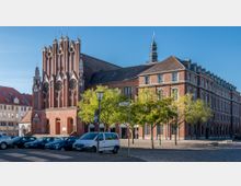 Backstein-Gebäudeensemble mit einer Kirchenfassade im gotischen Stil links und einem größeren, angrenzenden Backsteingebäude rechts, umgeben von Bäumen, parkenden Autos und einer Kopfsteinpflasterstraße. Im Vordergrund steht eine Straßenlaterne, und der Himmel ist klar.