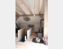 Wohnraum in einem Dachgeschoss mit sichtbaren Holz-Deckenbalken, weiß verputzten Wänden und Holzboden. Ein Kaminofen mit Holzkorb steht auf einer schwarzen Unterlage, daneben ein Treppenabgang mit weißen Wänden, im Hintergrund Regalflächen hinter Vorhängen.